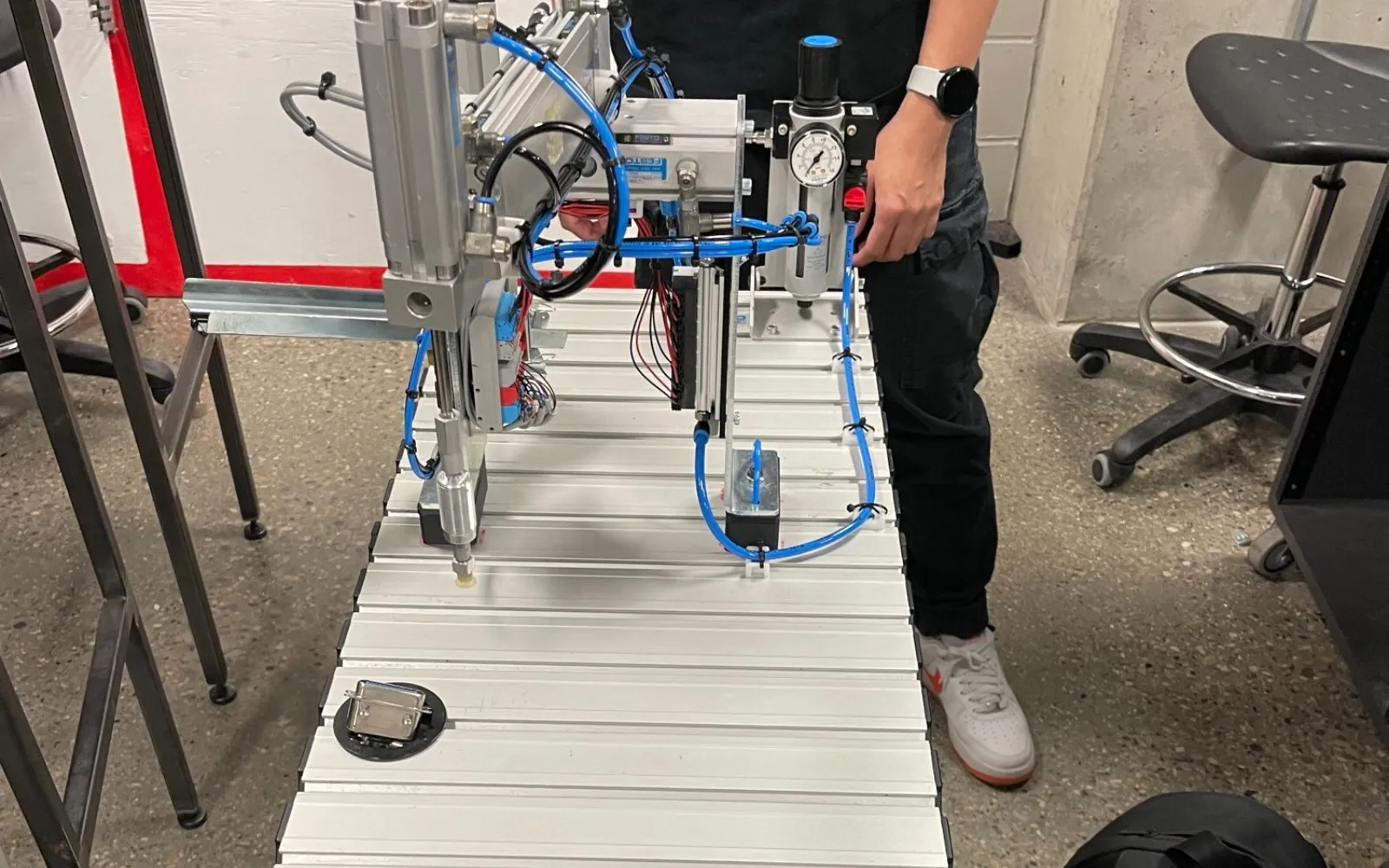 Festo MPS handling station with a pneumatic 3-axis arm, air service unit, and blue air lines on an aluminum work table.