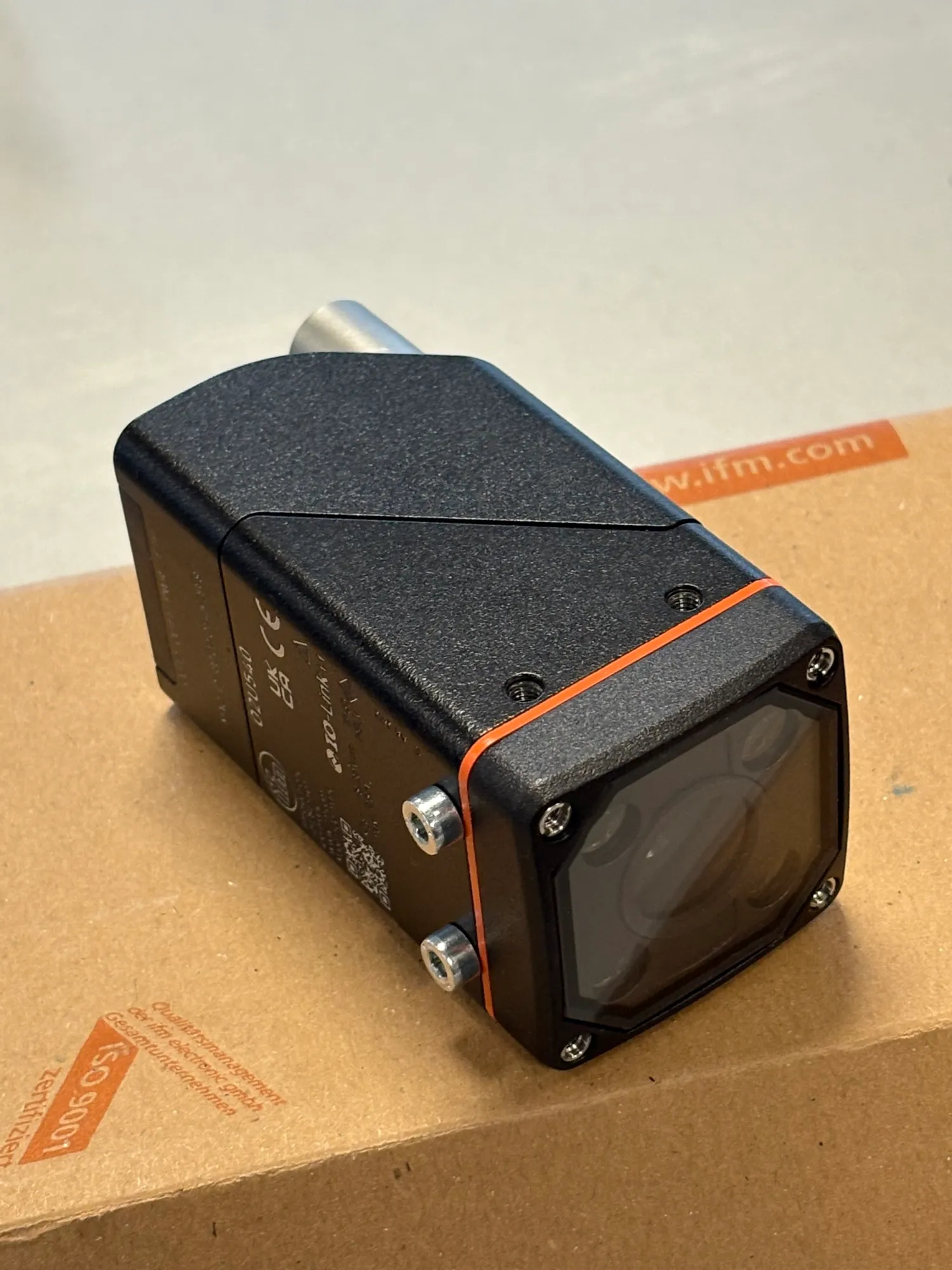 Close-up photo of the black IFM vision camera with an orange trim line, resting on its shipping box.